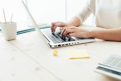 Woman typing on an Apple laptop on a clean desk with yellow sticky notes and a calculator and a pencil cup with 3 pencils