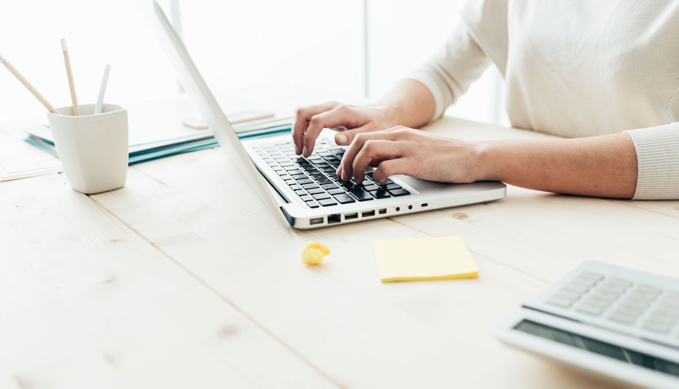 Woman typing on an Apple laptop on a clean desk with yellow sticky notes and a calculator and a pencil cup with 3 pencils