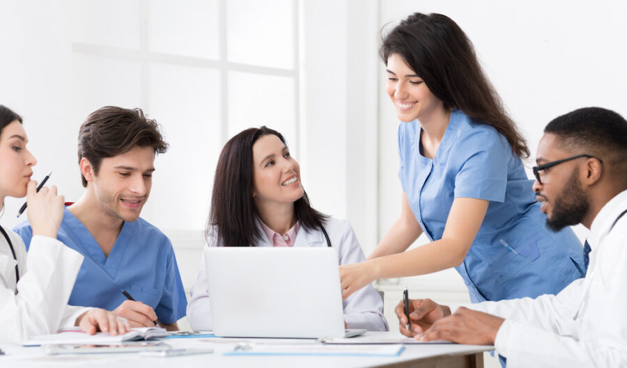 Medical personnel working together at a table. There is one person working on a laptop while others take notes in a notebook.