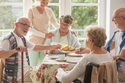 Group of four senior citizens eating a meal while a nurse stands beside their table